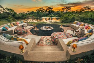 Un espacio circular al aire libre con sofás y cojines, una chimenea y vistas a un lago al atardecer.