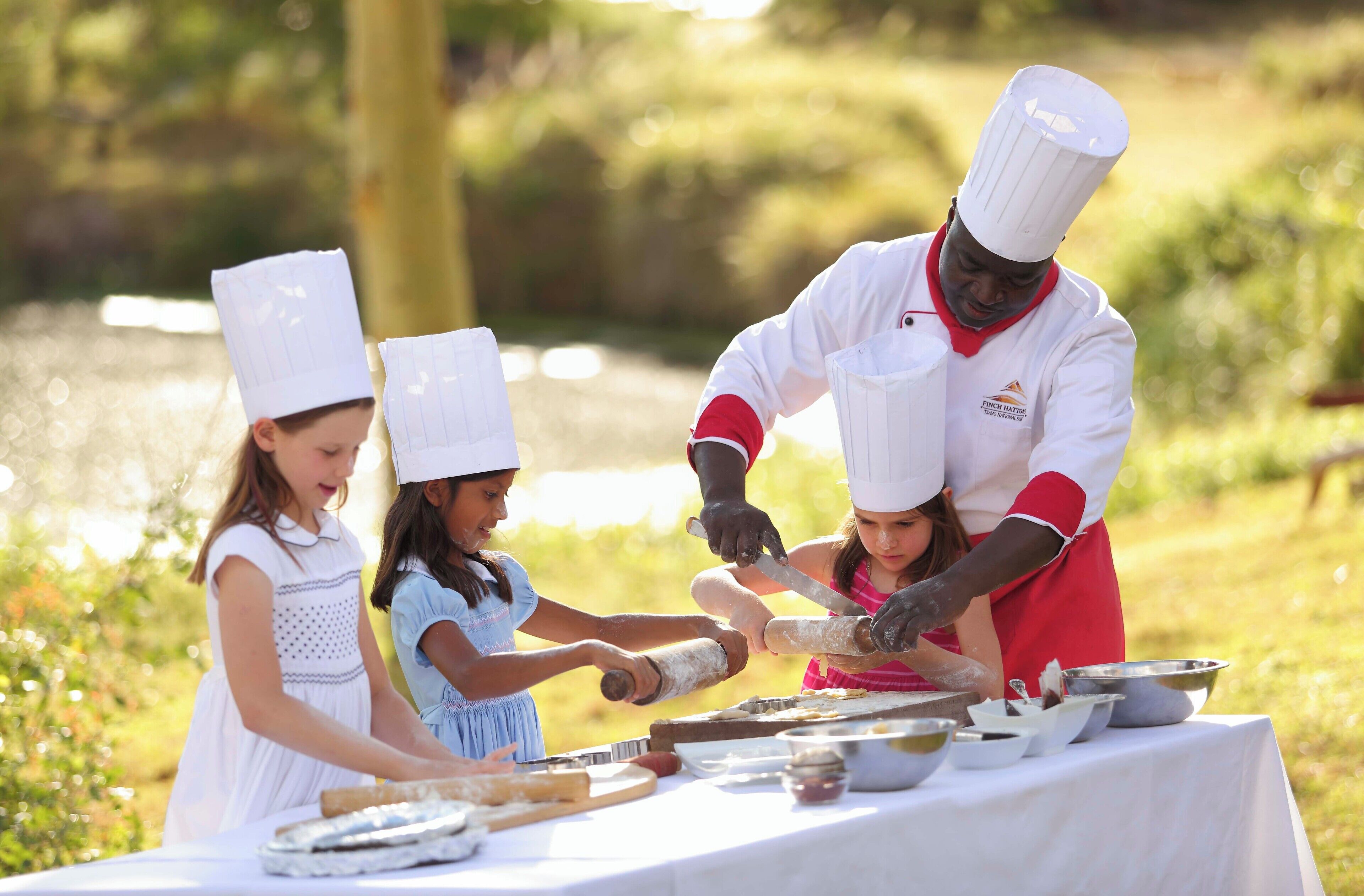A chef teaching children to roll dough at an outdoor table with a white tablecloth.