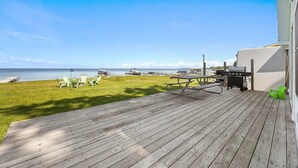 Una terraza de madera con una mesa de picnic y una parrilla, con vistas a una zona verde con sillas y una vista del agua.