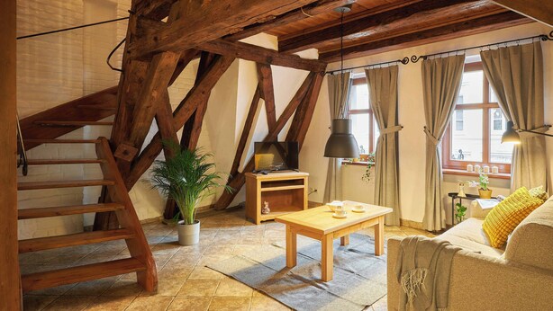 A rustic attic room with exposed wooden beams, a wooden table, a sofa, and a staircase. - Vincent Old Town Apartments  (Wroclaw)