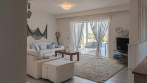 Una sala de estar con un sofá blanco, una mesa de centro y una televisión. Se ve un ventanal con cortinas y vistas al exterior.