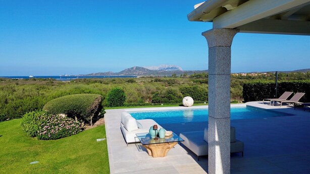 Un área de piscina con mesa de vidrio, sillas de descanso y vistas al mar y a las montañas.