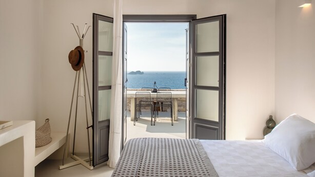A bedroom with a bed, a nightstand, a window with curtains, and a view of the sea and a table outside. - Ocean Villa - Res. I: Experience the Aegean Magnificence in style and privacy! (Paros)