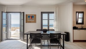 A dining area with a table and chairs, a view of the sea through open doors, and a kitchen area with cabinets and a window. - Ocean Villa - Res. I: Experience the Aegean Magnificence in style and privacy! (Paros)