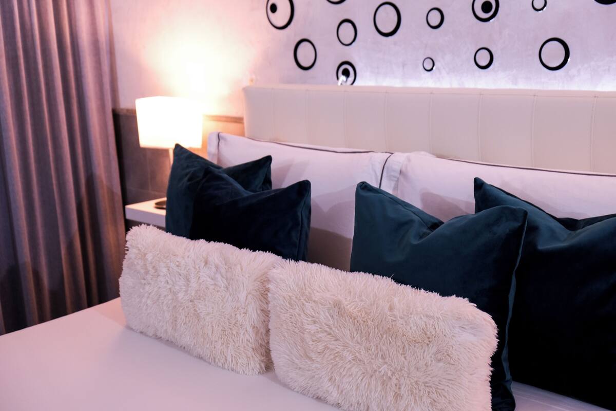 A hotel room with a white bed, fluffy pillows, a nightstand with a lamp, and a wall with circular patterns.