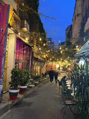 Una stretta via fiancheggiata da aree con tavoli all'aperto e vasi di piante, con luci decorative appese.
