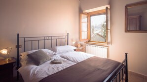 A neatly made bed with white linens and a brown blanket, a bedside table with a lamp, and a window with a view of trees and a building. - Sassocupo Apartment (Arezzo)