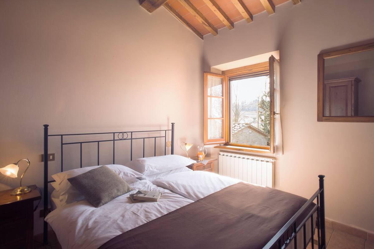 A neatly made bed with white linens and a brown blanket, a bedside table with a lamp, and a window with a view of trees and a building.