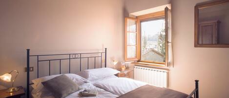 A neatly made bed with white linens and a brown blanket, a bedside table with a lamp, and a window with a view of trees and a building.