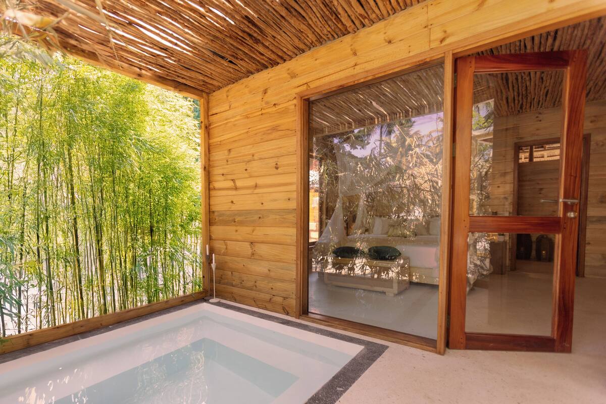 Una habitación de madera con un ventanal que da al exterior, un muro de bambú y un pequeño rincón de estar interior.