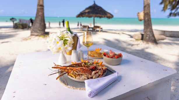 A table with a seafood dish, a glass of wine, and a bowl of fruit on a beach with a thatched umbrella and palm trees.