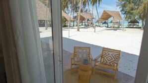 A beach view with thatched-roof structures, two wooden chairs with cushions, and a woven mat on the sand.