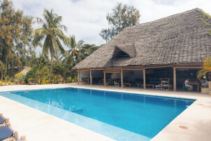 Zona de piscina con una estructura de techo de paja, sillas de descanso y palmeras.