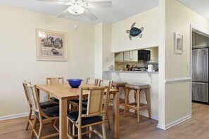 A dining area with a wooden table and chairs, a blue bowl, a ceiling fan, a kitchen with a refrigerator and microwave, and a framed picture on the wall.