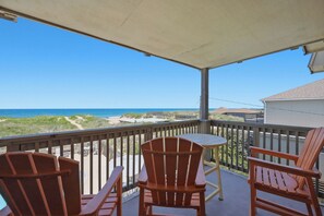 Ein überdachter Balkon mit Holzstühlen, Blick auf einen Strand mit Dünen und einem klaren Himmel.
