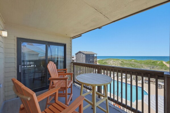A balcony with wooden chairs and a table, overlooking a swimming pool and beach.