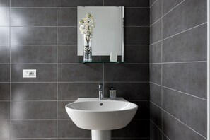 A bathroom with a white sink, dark tiled walls, a mirror, and a vase with flowers on a shelf.