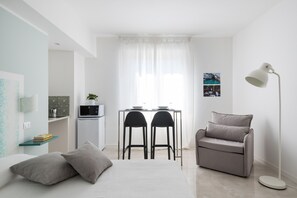 A compact, modern room with a bed, a desk with two chairs, a sofa, and a floor lamp.
