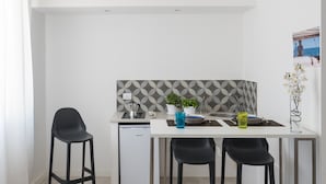 Et kompakt køkken med en hvid bordplade, indbygget ovn og et lille køleskab. To sorte stole, et blåt glas og en vase med blomster står på bordpladen.