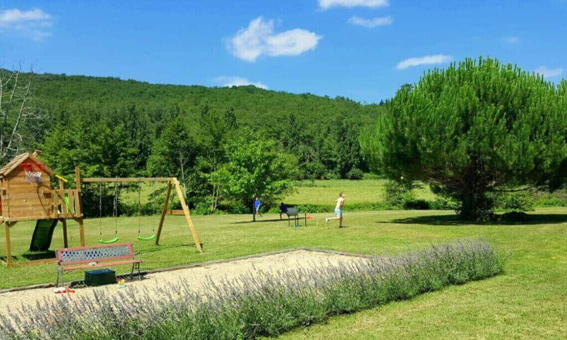 Une aire de jeux avec des balançoires et un toboggan, entourée d’un champ herbeux et d’une colline boisée.