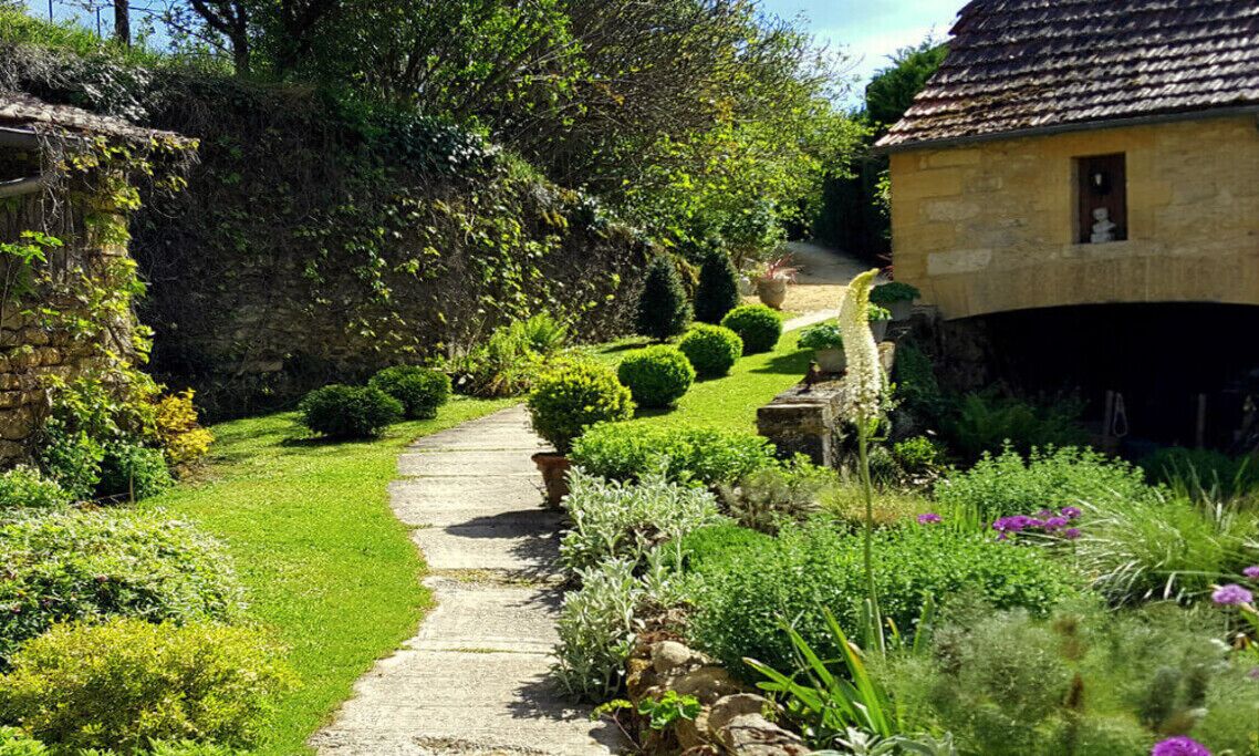 A stone pathway leading through a well-maintained garden with neatly trimmed hedges and a variety of plants.
