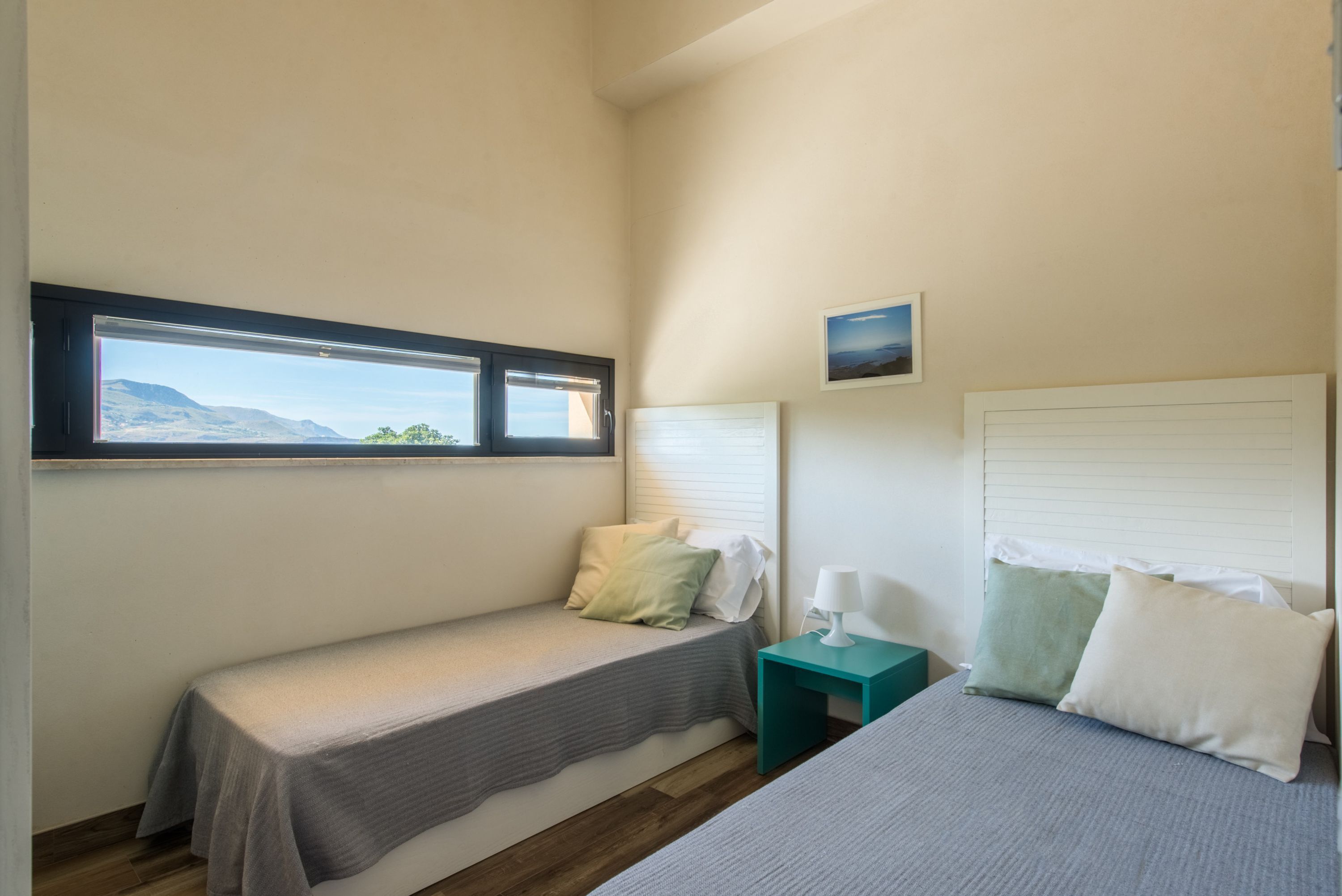Un dormitorio con una cama individual, una cama más pequeña, una ventana con vistas, una mesita de noche y un cuadro colgado en la pared.