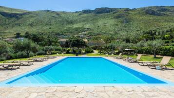Una piscina rectangular con baldosas de piedra, rodeada de sillones de descanso y follaje, con un paisaje montañoso al fondo.