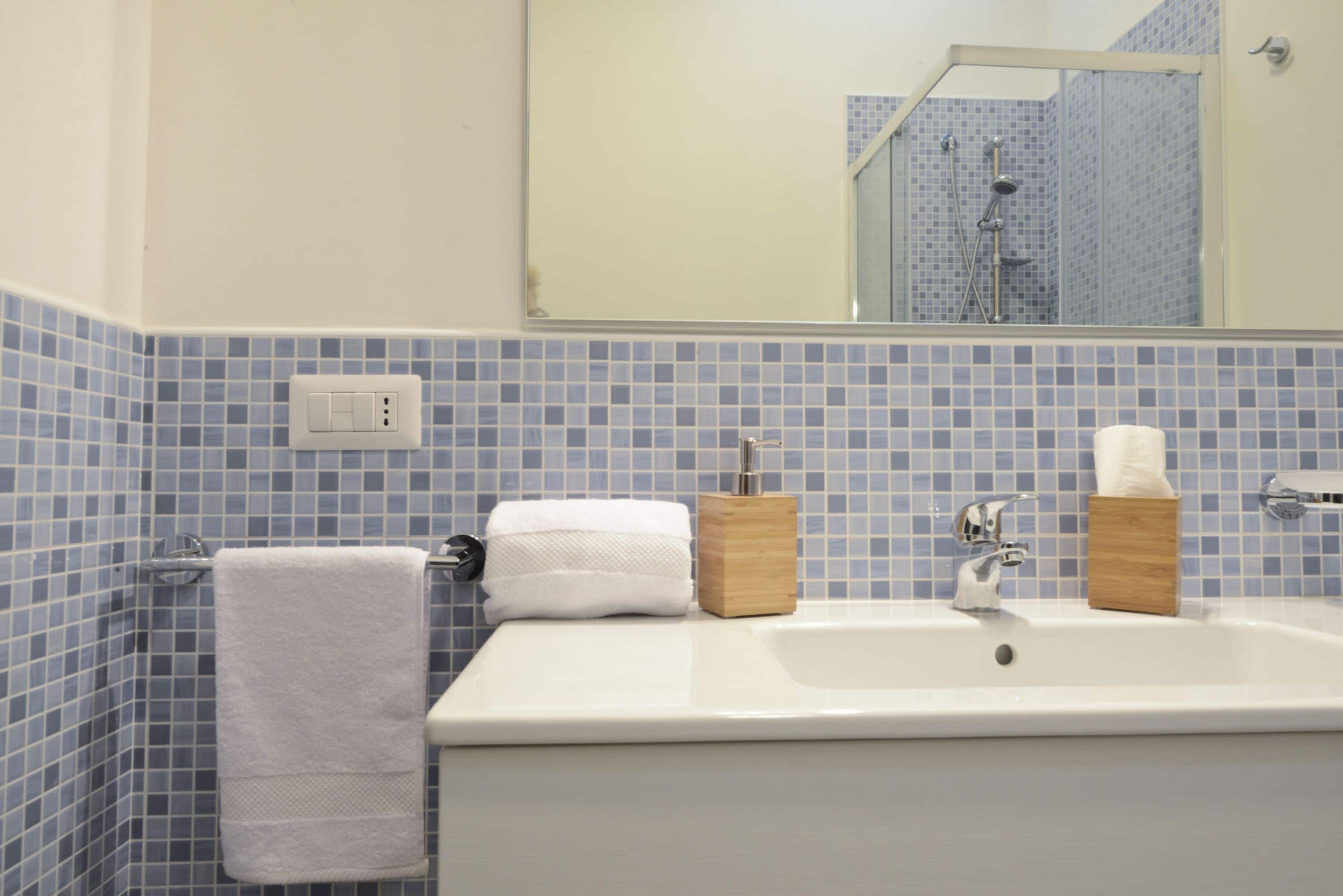 Ein Badezimmer mit einem weißen Waschbecken, blauen Mosaikfliesen, einem weißen Handtuch und einem hölzernen Seifenspender.