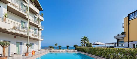 A swimming pool with lounge chairs and a view of the sea, surrounded by apartment buildings.