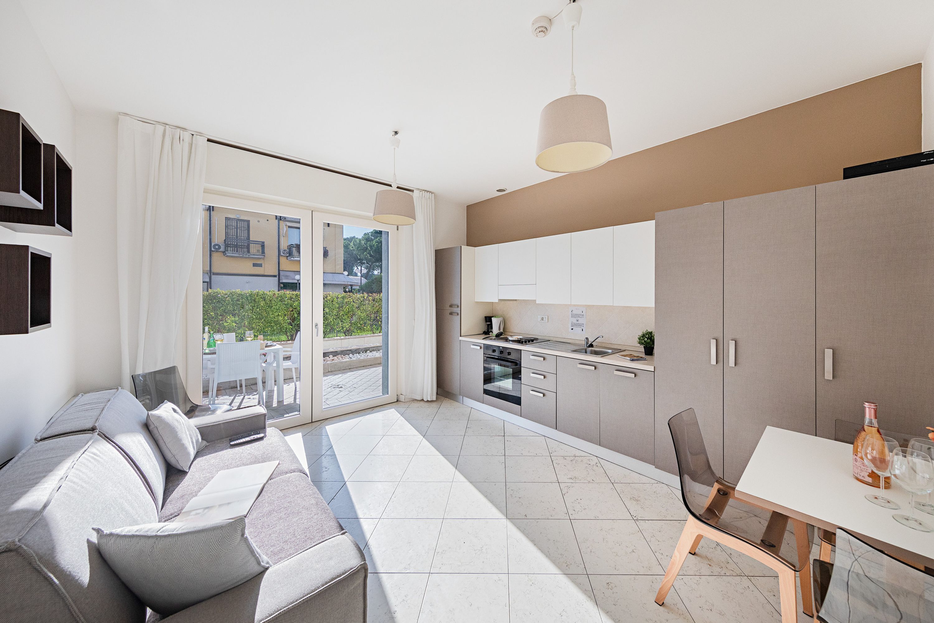 A modern living room with a sofa, a dining table with chairs, a kitchen area, and a sliding glass door leading to an outdoor patio.