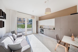 A modern living room with a sofa, a dining table with chairs, a kitchen area, and a sliding glass door leading to an outdoor patio. - Residenza Miralago - 04 Augusta - Locazione BK (Desenzano del Garda)