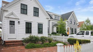 A two-story residential house with a white exterior, a brick front porch, and a white picket fence.