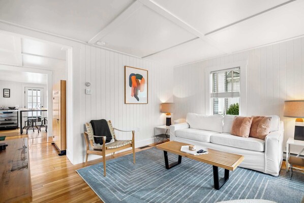 Un salon spacieux avec un canapé blanc, une table basse en bois et un fauteuil. Un tableau orne le mur, et une fenêtre est équipée de stores.