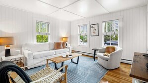 A modern living room with a white sofa, a wooden coffee table, and a patterned rug. There are two windows with blinds, a framed picture on the wall, and a lamp on a side table.