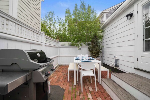 A backyard with a barbecue grill, a table set for four, and a white fence. - Stylish, Walk to Town and Beach! (Provincetown)