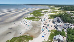 An aerial view of a beach with numerous umbrellas and people, a parking lot, and surrounding greenery.