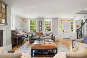 A spacious living room with a grand piano, a sofa, a coffee table, and large windows offering a view of greenery.