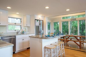 A modern kitchen with white cabinetry, a stainless steel refrigerator, and a dining area with wooden tables and chairs. - Waterfront on Follins Pond! (Dennis)