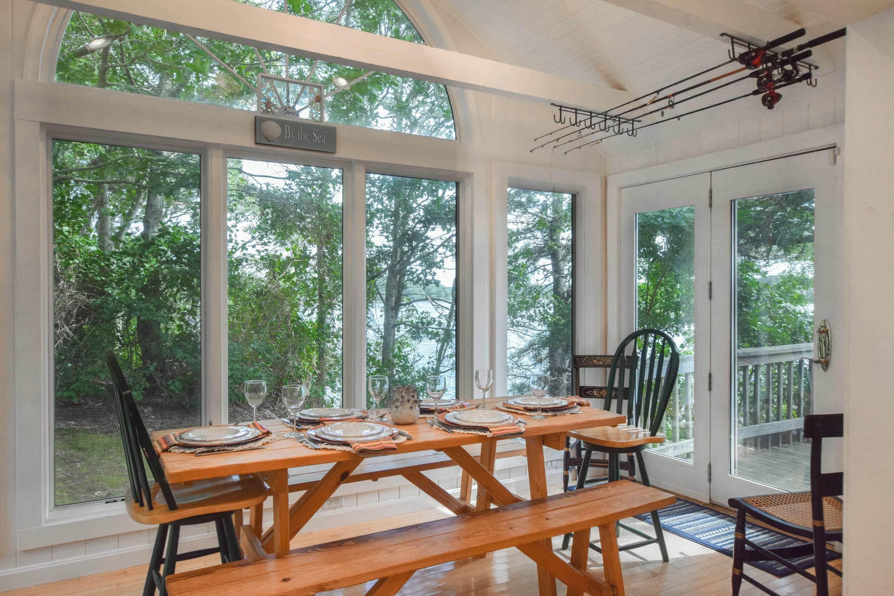 A dining area with a wooden table set for four, large windows offering a view of the trees, and a ceiling fan.
