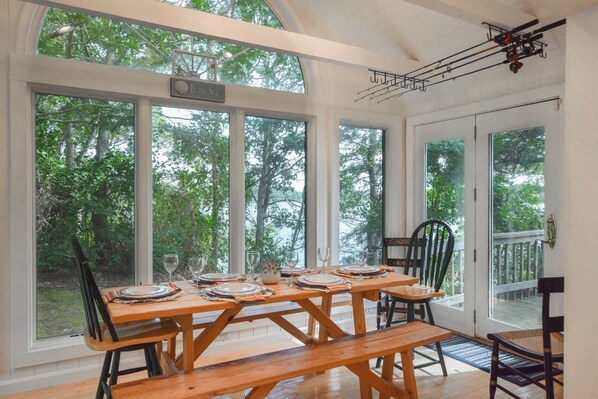 A dining area with a wooden table set for four, large windows offering a view of the trees, and a ceiling fan.