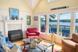A cozy living room with a fireplace, comfortable seating, a glass coffee table, and a view of the sea.