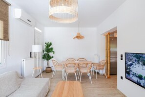 Ein modernes Wohnzimmer mit einer Couch, einem Esstisch, Stühlen, einer Hängelampe, einer Zimmerpflanze und einem Fernseher an der Wand.