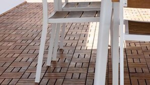 Una mesa blanca y dos sillas de madera sobre un piso de madera con diseños, con una pared blanca y una ventana al fondo.