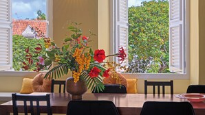 Un coin repas comprenant une table, des chaises, un vase de fleurs et une vue sur une maison par de grandes fenêtres ouvertes.