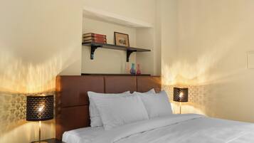A neatly made bed with white linens, two bedside lamps, a shelf above the bed with books and decorative items, and a headboard.