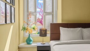 A bedroom with a bed, a nightstand with a lamp, a vase with flowers, and a window with shutters. - Drift Hotels  (Willemstad)