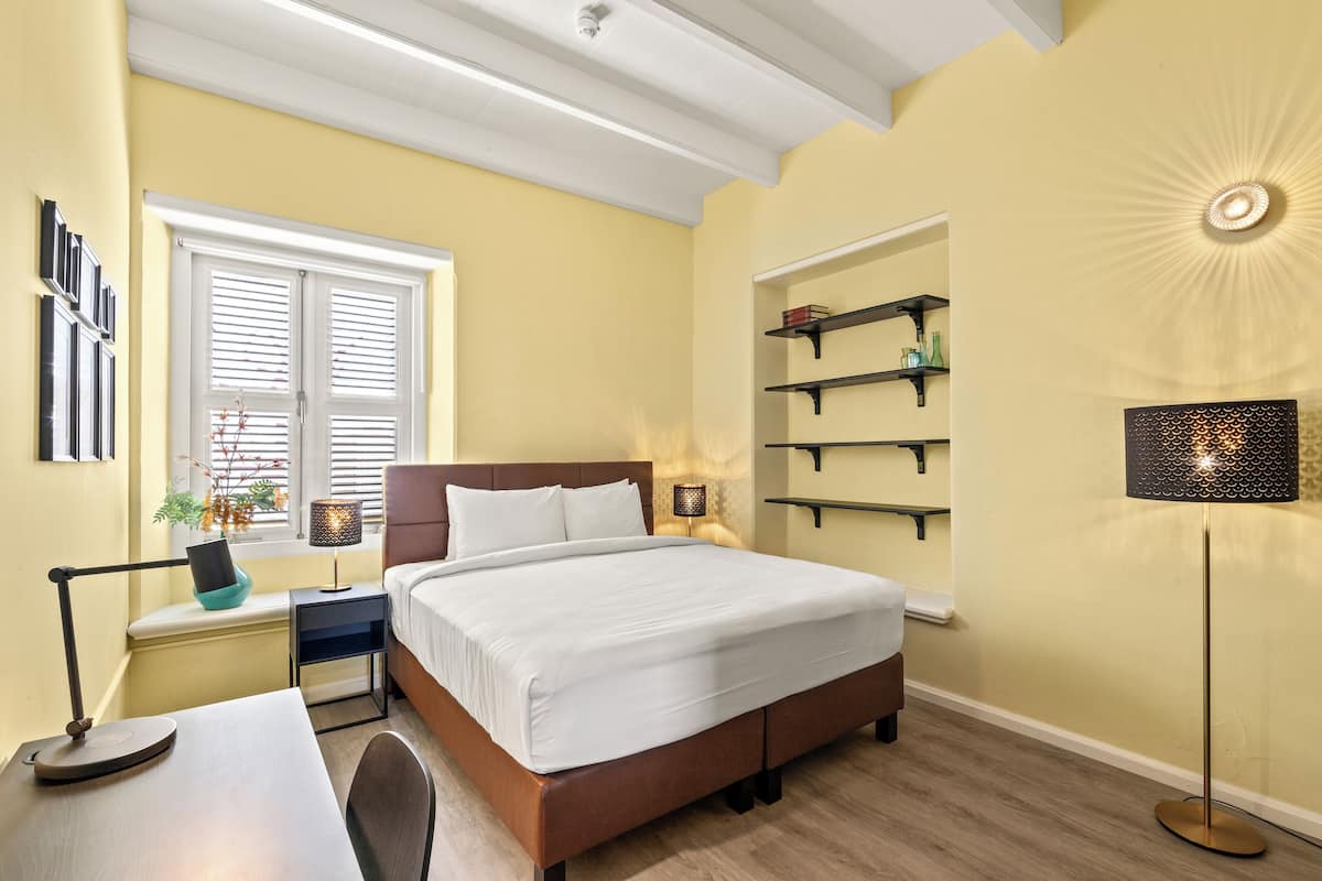 A bedroom with a bed, bedside tables, a desk, a window with blinds, shelves, and a floor lamp.