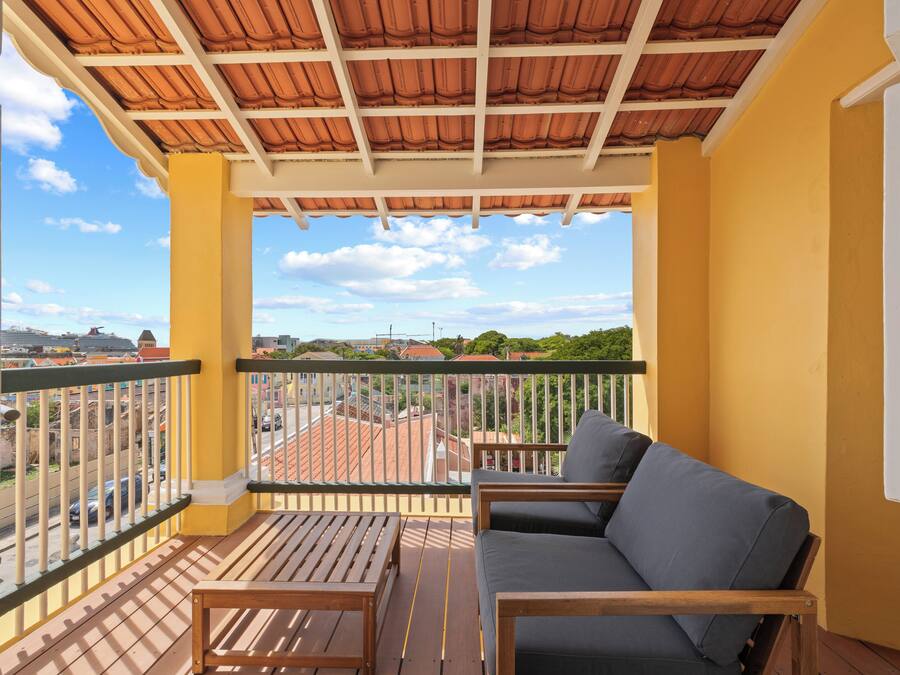 Een balkon met een houten tafel en een bank, gele muren, een pannendak en uitzicht op de stad.