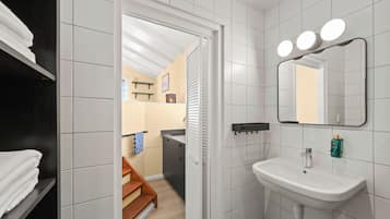 Un baño con lavamanos blanco, espejo y tres luces encima. Hay un perchero y un estante con artículos de aseo.