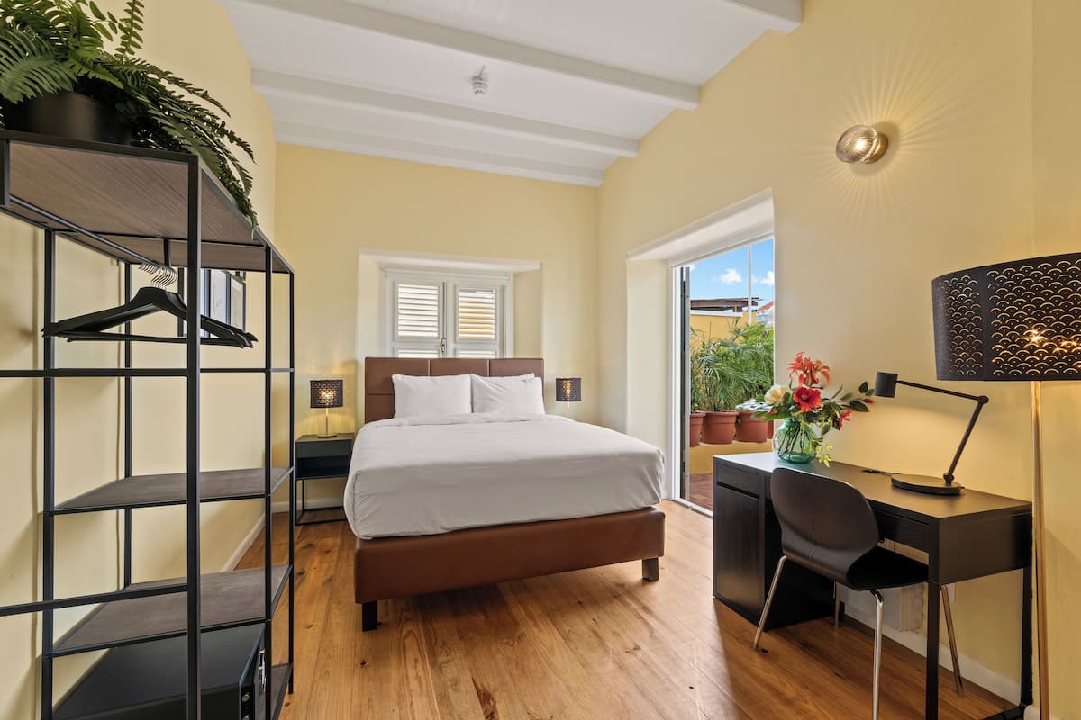 A bedroom with a bed, a desk, a chair, a shelf with clothes, a lamp, and a window.